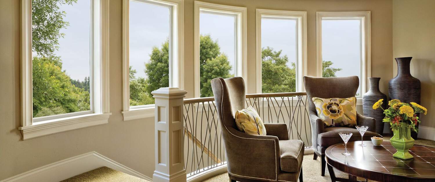Amazing view of trees outside of the Big 5 panel casement high efficient vinyl windows installed by a general contractor in a Light warm gray painted walls in foyer with a couple brown lounge chairs with yellow -black flower pattern fabric pillows on each chair next to a round coffee table in the middle, with two martini glasses and green apple color porcelain base with yellow flowers on top, and two very large brown floor baseson the back corner of the rails for the stairwell to access the lower floor Lo's Contracting Window Interior and Exterior Painting
