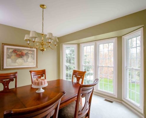 White Large Bay Vinyl Double-Hung Windows with white trimming on a light olive painted spacious elegant dining room decorated with golden 5 arm chandelier, gold framed painting showing ceramic bases with red flowers. Elegant hardwood table and chairs in the middle of the room. Sliding door installation, Patio door installation, Front door installation, windows and doors installation, energy efficient windows and doors replacement